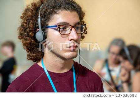 Berlin, germany, august 16, 2023. Young man with curly hair and glasses wearing headphones, intently listening to an audio guide while exploring a museum exhibition 137036598