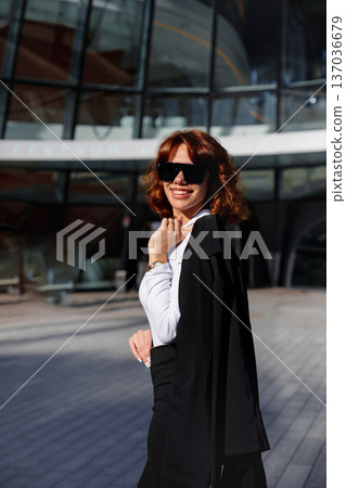 Smiling businesswoman in sunglasses standing near corporate building outdoors Smiling businesswoman in sunglasses standing near corporate building outdoors 137036679