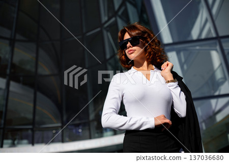 Elegant businesswoman holding jacket over shoulder near office building Elegant businesswoman holding jacket over shoulder near office building 137036680