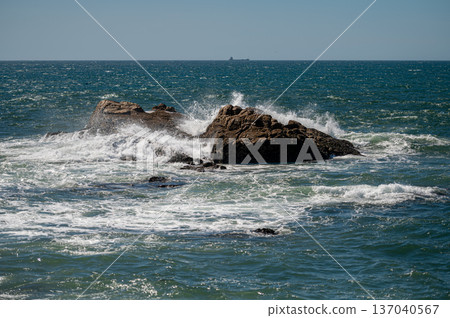 Waves crashing against a rocky shoreline with a distant cargo ship on a clear day Waves crashing against a rocky shoreline with a distant cargo ship on a clear day 137040567
