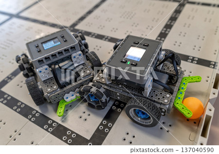 Two small robots are positioned on a grid-patterned floor, ready for a task involving a ball. Two small robots are positioned on a grid-patterned floor, ready for a task involving a ball. 137040590