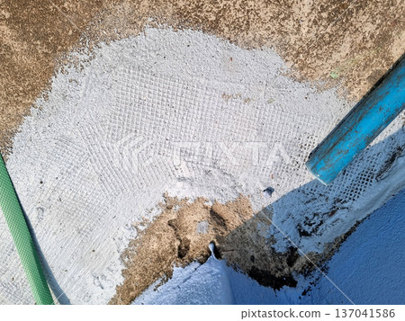 Professional waterproofing application on concrete surface. Features light gray sealant with reinforcement mesh texture over weathered material. Includes blue pipe and green construction strap. Professional waterproofing application on concrete surface. Features light gray sealant with reinforcement mesh texture over weathered material. Includes blue pipe and green construction strap. 137041586