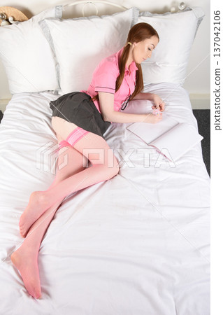 Beautiful redheaded model dressed as a schoolgirl in a pink shirt and grey pleated skirt, on her bed 137042106