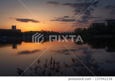 An evening city landscape of Minsk at sunset, reflected in a lakeside. Modern buildings and lights shimmer under a colorful sky. An evening city landscape of Minsk at sunset, reflected in a lakeside. Modern buildings and lights shimmer under a colorful sky. 137042238