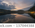 A calm lake at sunset on an autumn evening. Warm light from the colorful sky illuminates golden grass along the shore and a vibrant wild forest. 137044388