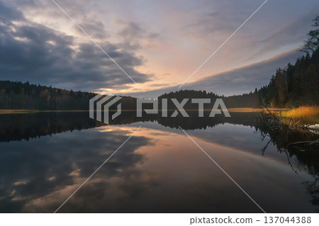 A calm lake at sunset on an autumn evening. Warm light from the colorful sky illuminates golden grass along the shore and a vibrant wild forest. A calm lake at sunset on an autumn evening. Warm light from the colorful sky illuminates golden grass along the shore and a vibrant wild forest. 137044388