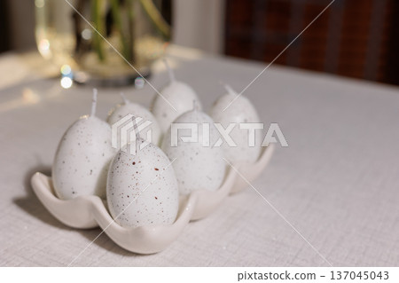 White speckled egg shaped candles standing in a cream ceramic holder on a white table, providing festive decor for an easter celebration or spring gathering White speckled egg shaped candles standing in a cream ceramic holder on a white table, providing festive decor for an easter celebration or spring gathering 137045043