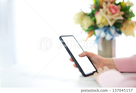 Hands of woman in pink shirt holding smart phone with white screen on white table top decorated with bouquet of flowers in a bright room background Hands of woman in pink shirt holding smart phone with white screen on white table top decorated with bouquet of flowers in a bright room background 137045157