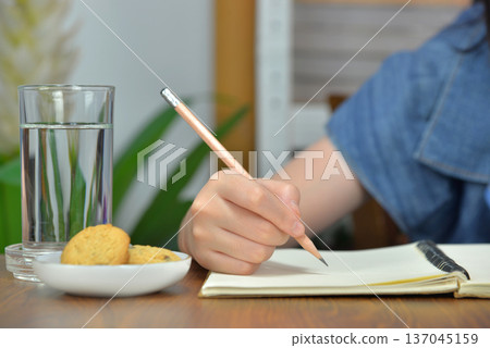 Woman's hand writing on notebook or doing homework on the wooden table at home There are a cookie and a glass of water on the side. 137045159