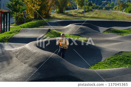Focused boy riding scooter through curved asphalt pump track in urban park. Active childhood, balance training,outdoor recreation and sporty lifestyle 137045365