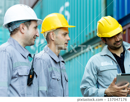 Construction workers wearing safety helmets discussing project details at shipping container yard, industrial teamwork and site coordination concept with tablet technology Construction workers wearing safety helmets discussing project details at shipping container yard, industrial teamwork and site coordination concept with tablet technology 137047176
