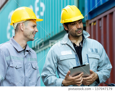 Two construction workers wearing yellow safety helmets discussing project details with digital tablet at shipping container yard, industrial teamwork and logistics management concept Two construction workers wearing yellow safety helmets discussing project details with digital tablet at shipping container yard, industrial teamwork and logistics management concept 137047179