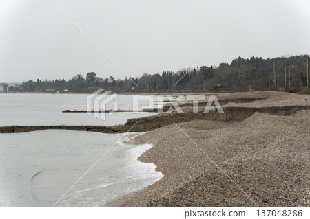 Pebble beach along calm coastline, soft waves, muted sky and distant tree line forming tranquil seaside panorama. Visual supports tourism promotion, coastal branding, sustainability themes Pebble beach along calm coastline, soft waves, muted sky and distant tree line forming tranquil seaside panorama. Visual supports tourism promotion, coastal branding, sustainability themes 137048286