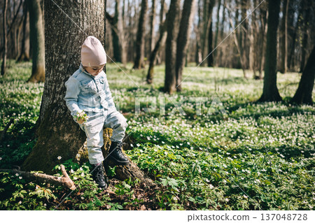 Little girl in jeans jacket standing near the tree in the forest covered with anemones Little girl in jeans jacket standing near the tree in the forest covered with anemones 137048728