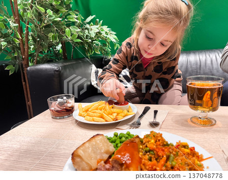 Child enjoys fries with ketchup at a diner while food is served on the table Child enjoys fries with ketchup at a diner while food is served on the table 137048778