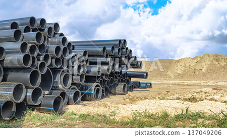 At a construction site surrounded by bare earth and grass, under a bright sky, black pipes are neatly stacked for a future water supply project 137049206