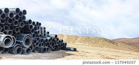 Black pipes stand tall in neat stacks on a sandy terrain, surrounded by hills as clouds drift overhead, capturing a moment of industrial stillness Black pipes stand tall in neat stacks on a sandy terrain, surrounded by hills as clouds drift overhead, capturing a moment of industrial stillness 137049207