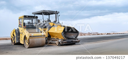 Heavy equipment-a roller and paver-works smoothly, laying asphalt, while construction crews lay the foundation for a new road against a picturesque cloudy backdrop Heavy equipment-a roller and paver-works smoothly, laying asphalt, while construction crews lay the foundation for a new road against a picturesque cloudy backdrop 137049217