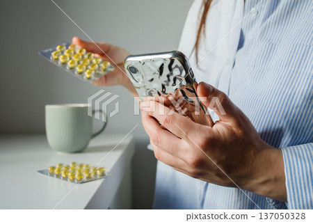 Woman taking fish oil omega 3 capsules for woman health and reading instruction for medication in the internet on smart phone close up. Supplements and vitamins, self treatment. 137050328