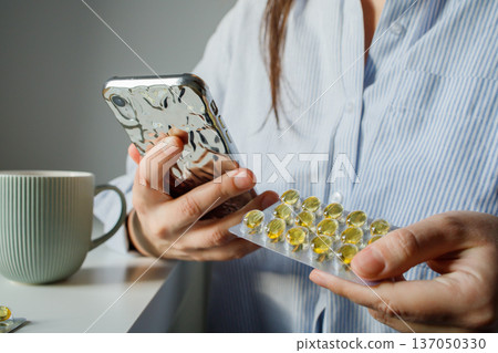 Woman taking fish oil omega 3 capsules for woman health and reading instruction for medication in the internet on smart phone close up. Supplements and vitamins, self treatment. Woman taking fish oil omega 3 capsules for woman health and reading instruction for medication in the internet on smart phone close up. Supplements and vitamins, self treatment. 137050330