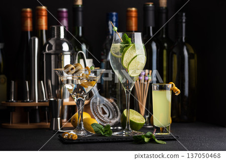 Bar composition featuring a variety of cocktails: martini with olives, citrus mint highball, and limoncello shot against wine bottles, shaker, strainer and fresh ingredients Bar composition featuring a variety of cocktails: martini with olives, citrus mint highball, and limoncello shot against wine bottles, shaker, strainer and fresh ingredients 137050468
