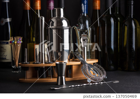 Professional cocktail bar composition featuring a stainless steel shaker, jigger, strainer, muddler, zester, and pour spouts neatly arranged on a wooden stand against wine bottles in background Professional cocktail bar composition featuring a stainless steel shaker, jigger, strainer, muddler, zester, and pour spouts neatly arranged on a wooden stand against wine bottles in background 137050469