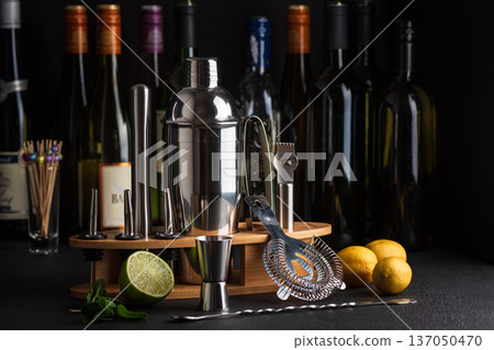 Professional cocktail bar composition featuring a stainless steel shaker, jigger, strainer, muddler, zester, and pour spouts neatly arranged on a wooden stand against wine bottles in background 137050470