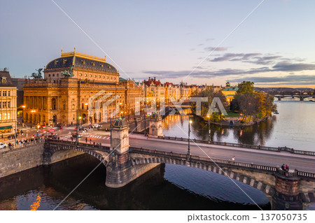 Aerial view of the National Theatre in Prague during the evening. The theatre is lit up, and the surrounding embankment and bridges are visible. Aerial view of the National Theatre in Prague during the evening. The theatre is lit up, and the surrounding embankment and bridges are visible. 137050735