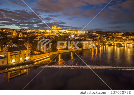 Aerial view of Prague at night shows illuminated Prague Castle and Charles Bridge over the Vltava River. The lights reflect on the water and the cityscape. 137050758