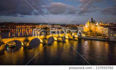 Charles Bridge stands illuminated at night in Prague. The river reflects the lights of the surrounding buildings, creating a vibrant scene by the embankment during the evening. 137050760