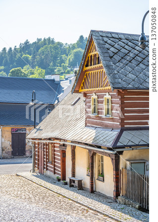 Timbered houses stand in a village conservation area in Zelezny Brod-Travniky. Cobblestone streets lead to the buildings under clear skies. Trees surround the area providing greenery. 137050848
