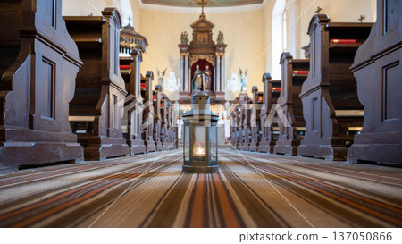 A lantern holding the Bethlehem light stands on the floor of a church. The surrounding wooden pews create a simple scene of worship. Soft light shines from the lantern. A lantern holding the Bethlehem light stands on the floor of a church. The surrounding wooden pews create a simple scene of worship. Soft light shines from the lantern. 137050866