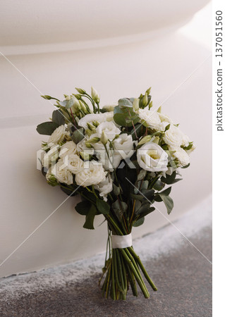 White carnation wed bouquet with wrapped stems leaning against white wall in sunlight 137051560