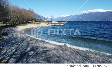 Pebble beach in Toscolano Maderno on Lake Garda with snow-capped mountains. Pebble beach in Toscolano Maderno on Lake Garda with snow-capped mountains. 137051872