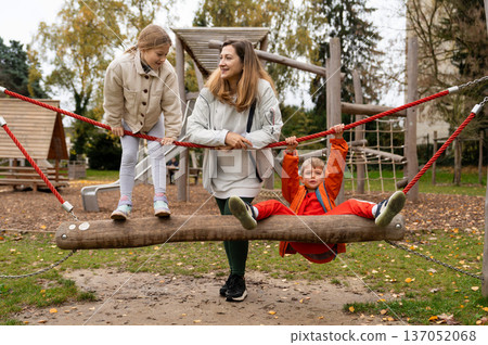 Mother guiding kids balancing on wooden playground rope bridge swing 137052068