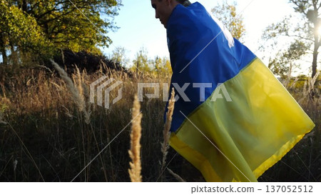 Young male military in uniform going with blue-yellow banner on shoulders at countryside. Soldier of ukrainian army walking with national flag as symbol of victory against russian aggression 137052512