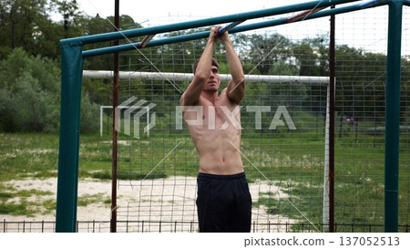 Young muscular sportsman doing pull ups at sports ground. Athletic guy exercising outdoor. Strong sporty man working out at summer nature. Concept of healthy and active lifestyle. Slow motion 137052513