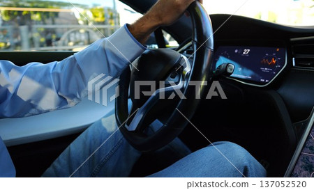 Male hand of businessman holds steering wheel while driving electrical vehicle at city. Businessperson operating electric car on urban road. Young man commuting to work at sunny day. Close up Male hand of businessman holds steering wheel while driving electrical vehicle at city. Businessperson operating electric car on urban road. Young man commuting to work at sunny day. Close up 137052520