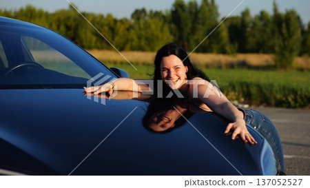Beautiful smiling girl bought new electrical vehicle and hugging hood auto. Happy young woman enjoying modern electric automobile. Concept of buy or rent car. Slow motion 137052527