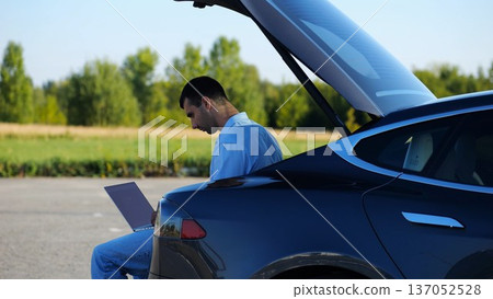Young handsome entrepreneur works on notebook sitting in electric car trunk. Successful businessman in formal wear using computer at auto. Confident manager typing text on laptop outdoor. Slow mo 137052528