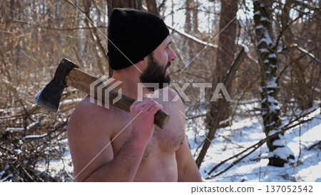 Portrait of shirtless bearded man stands in a snowy winter forest with axe. Tired guy resting after chopping logs in snowy woodland. The scene represents outdoor work in cold conditions. Close up 137052542
