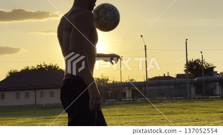 Young man juggling soccer ball on stadium at sunset. Professional footballer kicking ball at green field. Sportsman practicing tricks at meadow with sunlight at background. Freestyle football Young man juggling soccer ball on stadium at sunset. Professional footballer kicking ball at green field. Sportsman practicing tricks at meadow with sunlight at background. Freestyle football 137052554