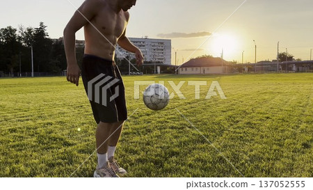 Young man juggling soccer ball on stadium at sunset. Professional footballer kicking ball at green field. Sportsman practicing tricks at meadow with sunlight at background. Freestyle football Young man juggling soccer ball on stadium at sunset. Professional footballer kicking ball at green field. Sportsman practicing tricks at meadow with sunlight at background. Freestyle football 137052555
