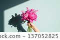A hand holds a beautiful, elegant bouquet of fresh flowers against a bright, light blue background. The arrangement features pink peonies, sweet peas 137052825
