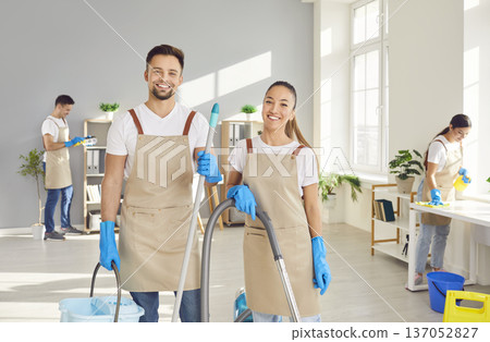 Young janitors looking at camera with a team of cleaning service washing office in background. 137052827