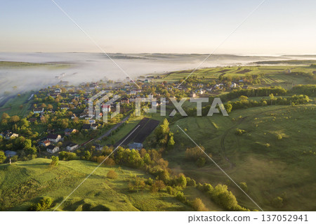 View from above of white fog on village house roofs among green trees under bright blue sky. Spring misty landscape panorama at dawn. View from above of white fog on village house roofs among green trees under bright blue sky. Spring misty landscape panorama at dawn. 137052941