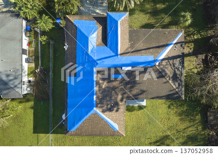 Aerial view of damaged in hurricane Ian house roof covered with blue protective tarp against rain water leaking until replacement of asphalt shingles Aerial view of damaged in hurricane Ian house roof covered with blue protective tarp against rain water leaking until replacement of asphalt shingles 137052958