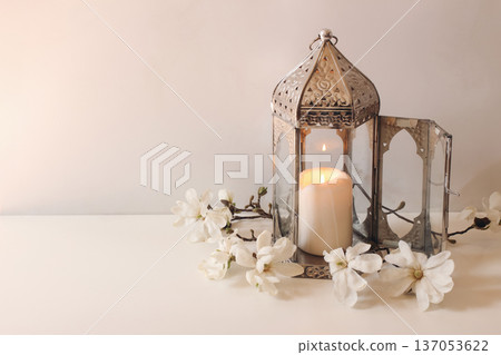 Ornamental silver Moroccan, Arab lantern with magnolia flowers on white table. Burning candle. Greeting card for holy month Ramadan Kareem. Eid mubarak celebration. Muslim religion banner. Ornamental silver Moroccan, Arab lantern with magnolia flowers on white table. Burning candle. Greeting card for holy month Ramadan Kareem. Eid mubarak celebration. Muslim religion banner. 137053622