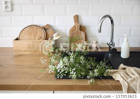 Black Kitchen Sink with Wooden Countertop, Cutting Boards and blurred fresh Cow Parsley Herbs Flowers. Modern Minimal Scandinavian Interior. White Metro Tiles Background, natural Light, Home Styling 137053623