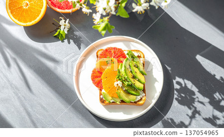 Spring avocado toast. Avocado toast with citrus slices on white plate, surrounded by flowers and fruit, in bright sunlight 137054965